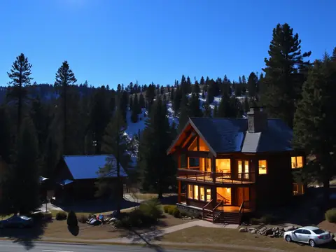 Two-story wood cabin with lit windows among tall pines at dusk
